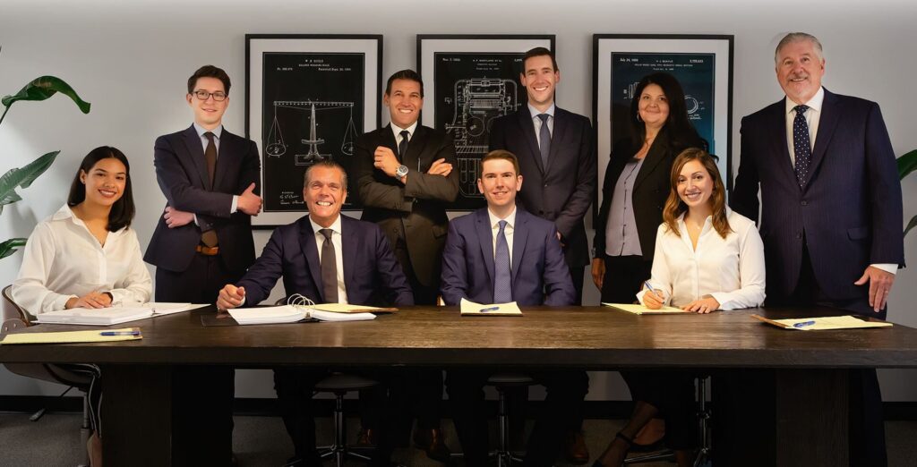 The legal team at Portale Randazzo LLP poses confidently in a professional office setting, with nine team members dressed in business attire, seated and standing in front of framed legal illustrations.