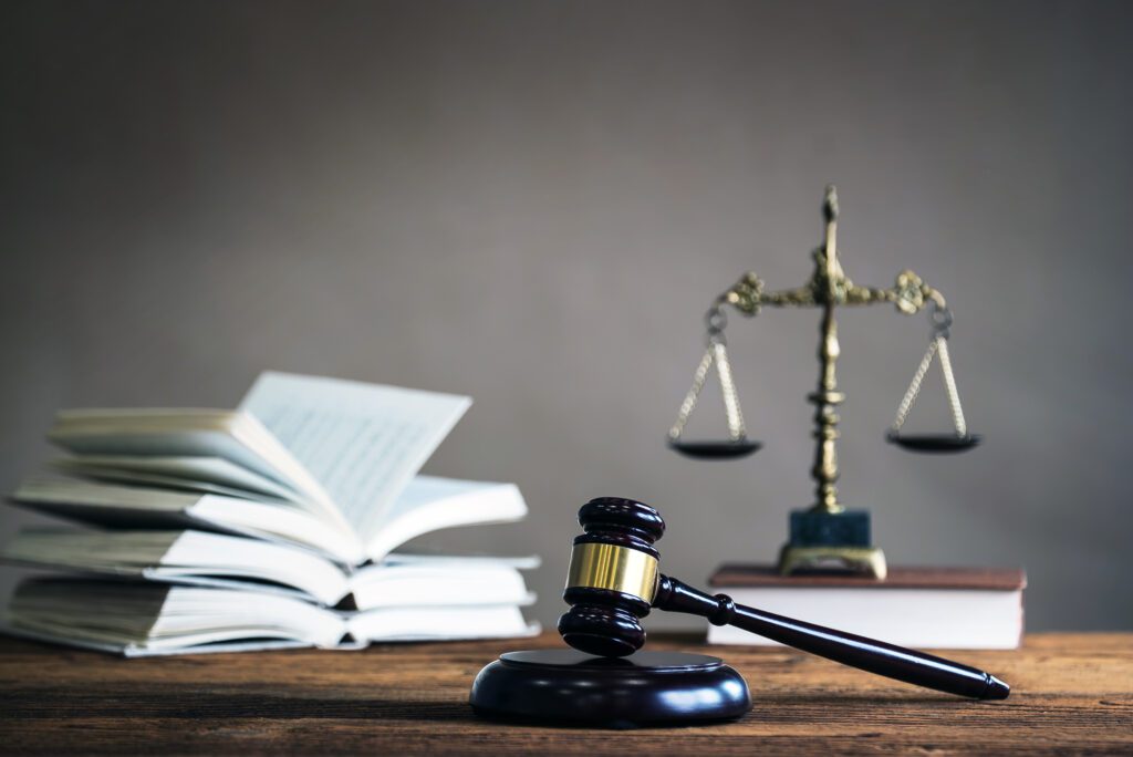 A judge’s gavel rests on a wooden desk beside a stack of open legal books, with a blurred brass scale of justice in the background, symbolizing law and justice.