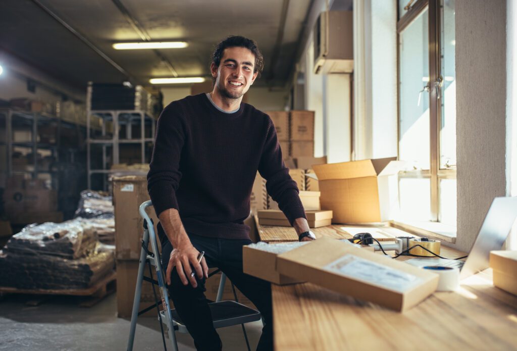 Smiling male ecommerce business owner in a fulfillment warehouse preparing online shopping orders for delivery, illustrating DTC brand logistics, inventory management, and customer experience.