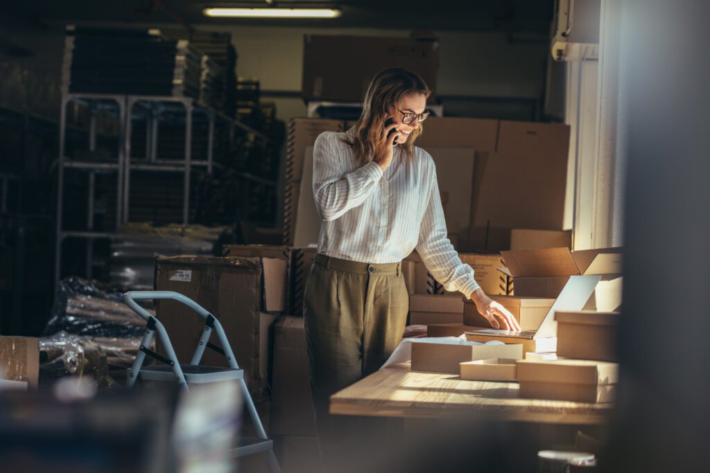 Female entrepreneur managing ecommerce order fulfillment in a warehouse while speaking on the phone, representing inventory operations, customer service, and ecommerce logistics for DTC brands.