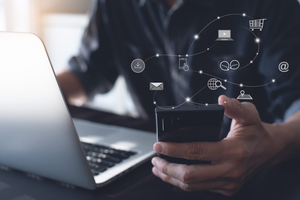 Close-up of a person using a smartphone and laptop with overlay icons for email marketing, ecommerce software, shopping cart, and social media, symbolizing omnichannel marketing automation and digital advertising tools for online retailers.