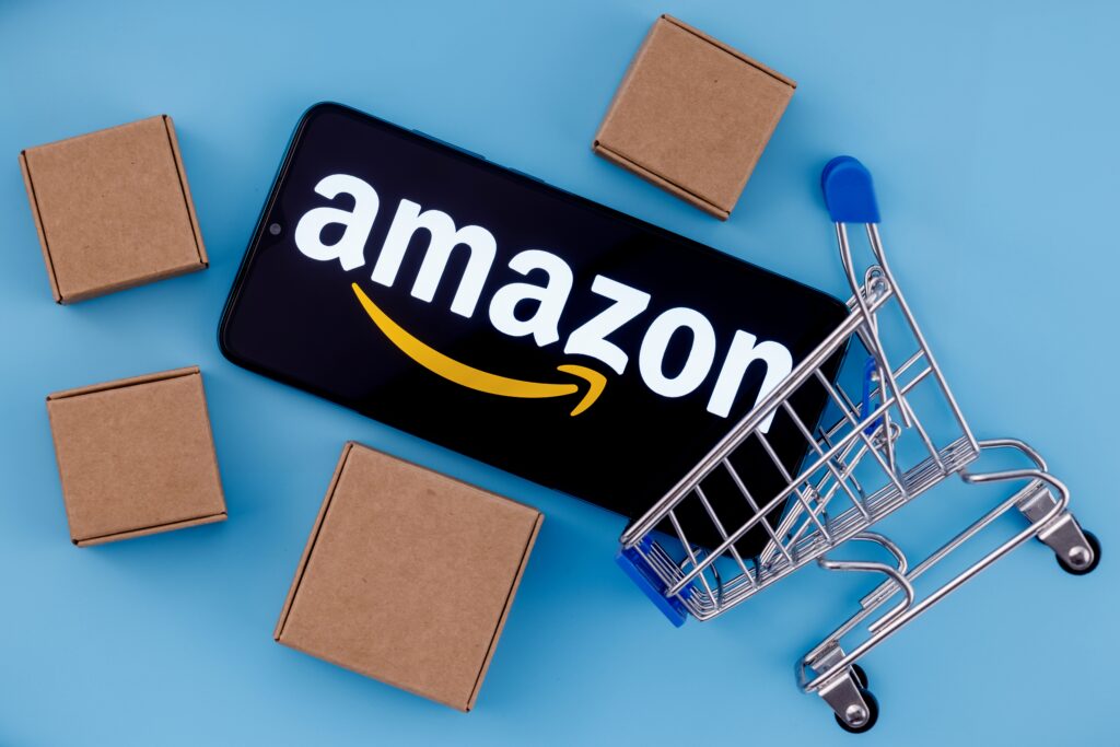 Smartphone displaying Amazon logo surrounded by cardboard boxes and a miniature shopping cart on a blue background, symbolizing ecommerce logistics, online shopping, Amazon marketplace, and product review-driven sales strategy.