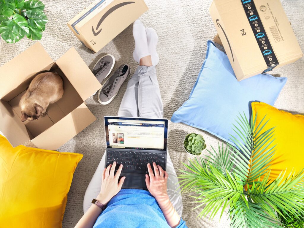 Overhead shot of person browsing Amazon on a laptop at home with delivery boxes, a cat in a box, and colorful pillows, representing consumer experience, online shopping, product research, and customer review engagement in ecommerce.
