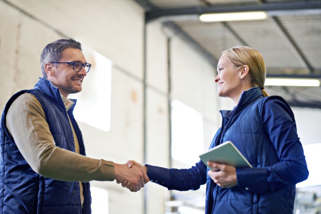 Business partners in a warehouse shaking hands after negotiating a B2B ecommerce contract focused on supply chain efficiency, product information management, and digital commerce scalability.