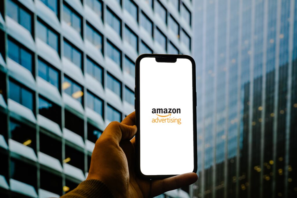 Amazon advertising displayed on smartphone held in front of office buildings, symbolizing ecommerce PPC strategy, digital marketing campaigns, and brand visibility growth in competitive online marketplaces.