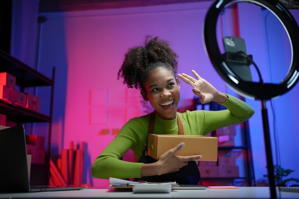Young woman filming UGC video with smartphone and ring light in ecommerce warehouse, showcasing influencer marketing, online shopping, and brand community engagement.