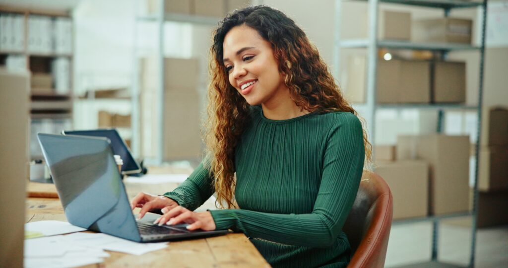 Smiling ecommerce business owner managing orders on a laptop in a warehouse, illustrating order fulfillment, inventory management software, and backend optimization for DTC SEO success.