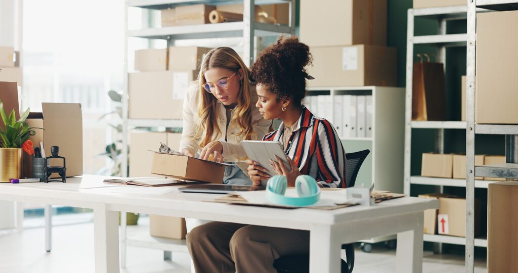 Ecommerce entrepreneurs managing product packaging and digital inventory on a tablet, symbolizing small business ecommerce, customer service, content strategy, and schema markup for better search engine results.