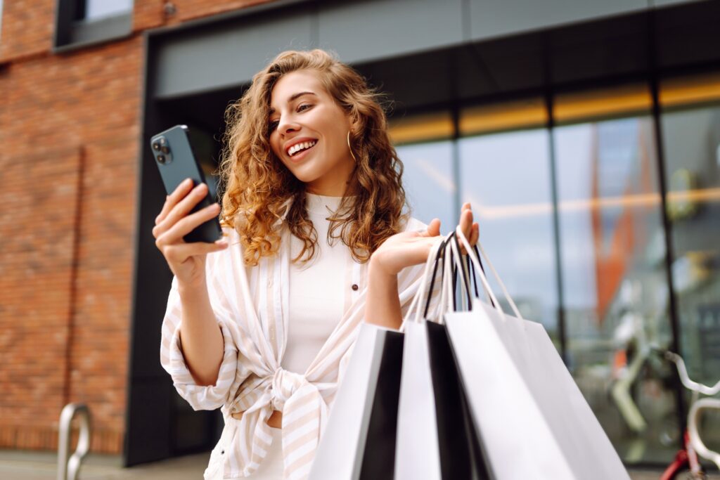 Happy shopper holding multiple retail shopping bags while using smartphone, illustrating omnichannel ecommerce, social commerce, and mobile shopping engagement.